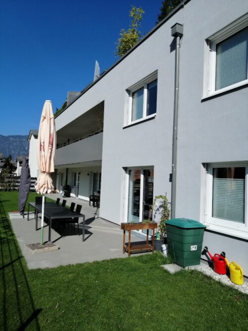 Terrasse eines Mehrfamilienhauses mit Esstisch, Stühlen, geschlossenem Sonnenschirm, Pflanzregal, grünem Abfallbehälter und zwei Gießkannen; Berge im Hintergrund; keine Personen sichtbar.