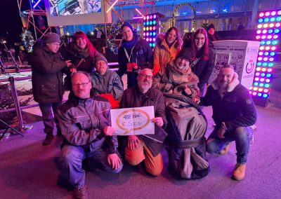 Mehrere Personen posieren vor einer Bühne mit Weihnachtsbeleuchtung und Ö3-Branding, halten ein Schild mit der Aufschrift „€ 500,30“; eine Spendendose/Box ist sichtbar.