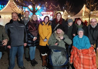 Mehrere Personen vor weihnachtlich beleuchteten Marktständen bei Nacht; im Hintergrund leuchtet die Schrift "HITRADIO Ö3"; vorn zwei Personen in Rollstühlen.