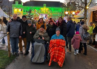 Gruppenfoto auf einem belebten Weihnachtsmarkt bei Dämmerung, vorn zwei Personen in Rollstühlen, im Hintergrund beleuchtetes Gebäude und Marktstände; auf einem Plakat ist „Weihnachtswunder“ sichtbar.