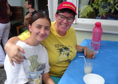 Zwei lächelnde Frauen sitzen an einem blauen Tisch mit Getränken; eine trägt ein rotes Cap mit der Aufschrift 'SAMARITERBUND' und ein gelbes T‑Shirt mit dem Schriftzug '10 Jahre'.