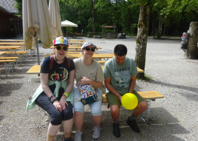 Drei Personen sitzen auf einer Holzbank im Außenbereich; Mann rechts hält einen gelben Ballon; auf seinem T‑Shirt steht 'SUMMER'.