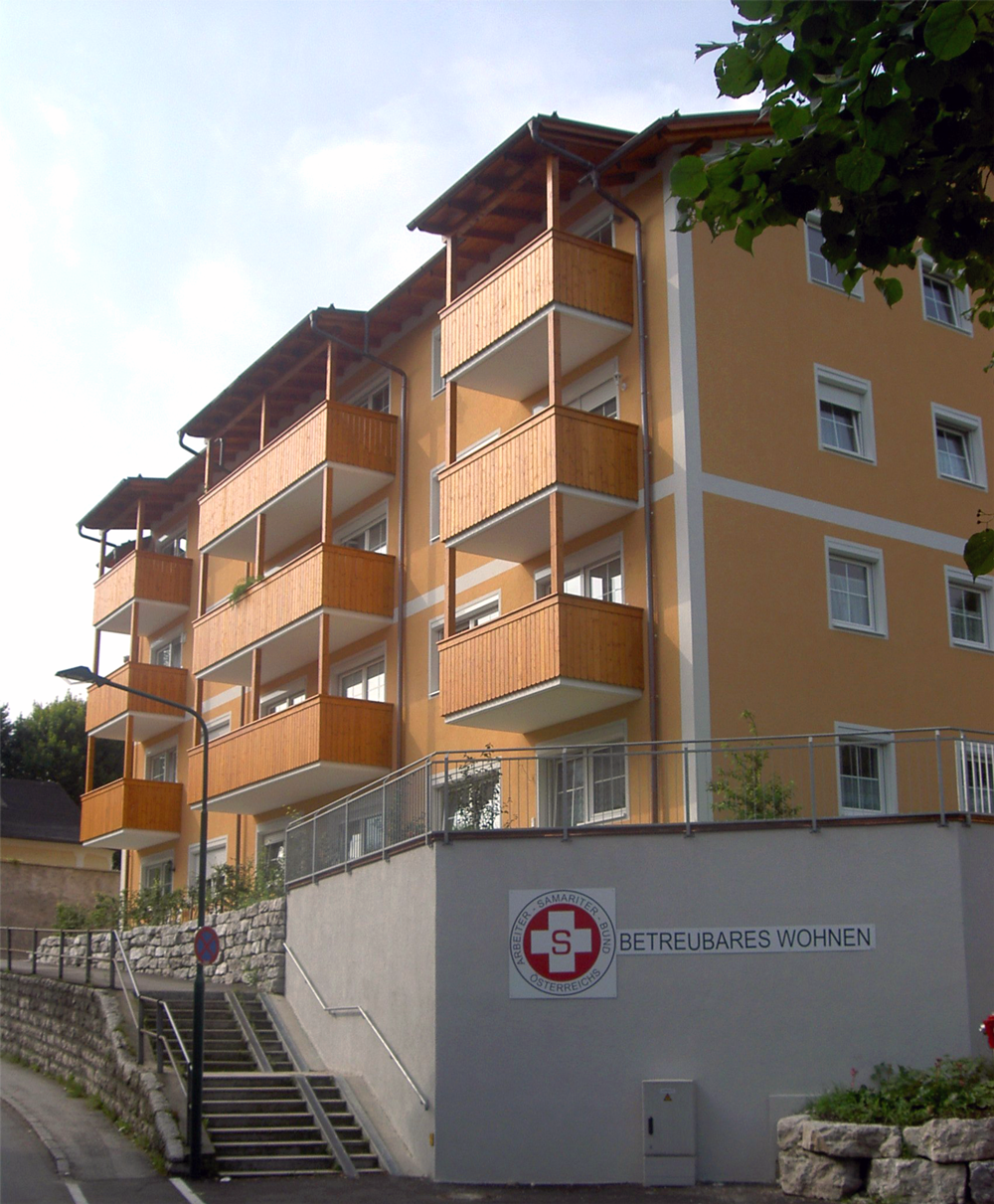 Mehrstöckiges gelbes Wohnhaus mit hölzernen Balkonen und Schild "BETREUBARES WOHNEN" an der Fassade.