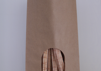 Braune Papiertasche mit Henkeln, ovales Sichtfenster zeigt Bündel dünner Holzstäbe, teils in netzartiger Umwicklung; kleines weißes Etikett unten rechts.