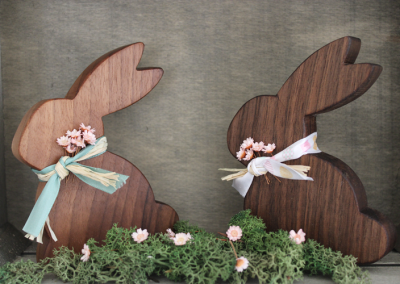 Zwei hölzerne Hasen-Silhouetten mit Stoffbändern und rosa Zierblumen auf Moos.