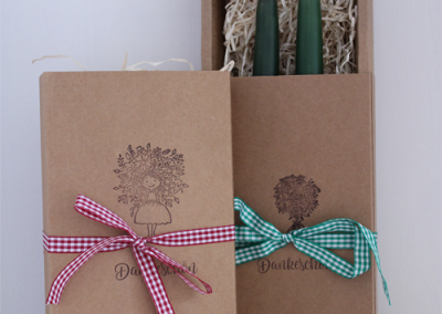 Zwei Kraft-Geschenkboxen mit Karobändern, offene Box mit Papier-Einstreu und zwei grünen Kerzen; Aufdruck "Dankeschön".