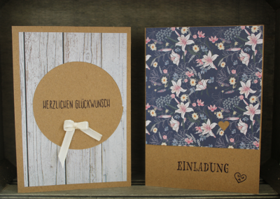 Zwei handgefertigte Karten auf einem Holzkasten; links Kraftpapierkarte mit rundem Element, Schleife und Text ‚HERZLICHEN GLÜCKWUNSCH‘; rechts Karte mit floralem Muster und Schrift ‚EINLADUNG‘.