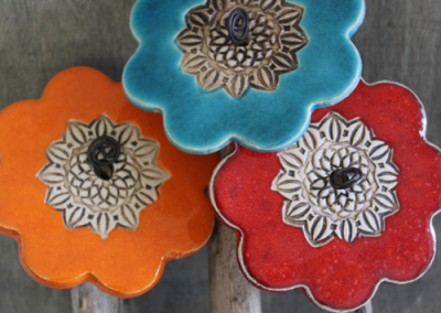 Drei blumenförmige Keramikornamente in Blau, Orange und Rot mit ornamentalen Metallzentren auf Holzstäben vor einer grauen Fläche.
