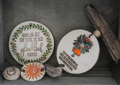 Drei handbemalte Tonscheiben in einem Holzfach; links mit dem Text „Nimm dir Zeit für Dinge, die dich glücklich machen“, rechts mit buntem Blumenkorb, unten eine Scheibe mit Sonne und Muschel