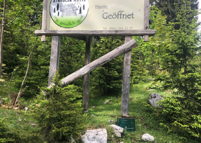 Holztafel am Waldrand mit Logo 'Ströbler Hütte', Pfeil '300 m' und dem Text 'Heute: Geöffnet'.