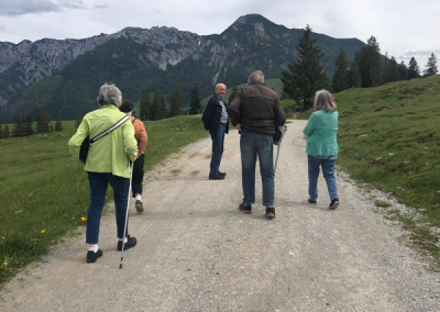 Fünf Personen von hinten auf einem Schotterweg, Spaziergang auf der Postalm vor Bergkulisse; zwei nutzen Wanderstöcke.