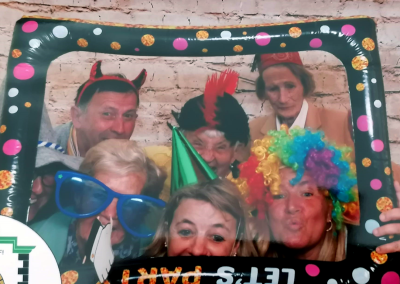 Gruppenbild im Fotobooth‑Stil vor einer Ziegelwand; Personen mit Kostümaccessoires (bunte Perücke, übergroße blaue Brille, Partyhüte, Hörner) halten einen gepunkteten, aufblasbaren Rahmen mit teilweisem Schriftzug "LET'S PARTY".