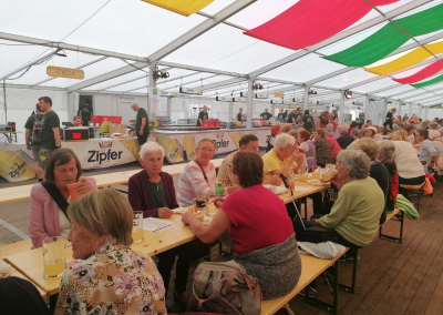 Innenansicht eines Festzelts mit Reihen von Bierbänken, zahlreichen Personen, die sitzen, essen und trinken; Ausschank mit "Zipfer"-Werbung und einem "Kassa"-Schild sichtbar.