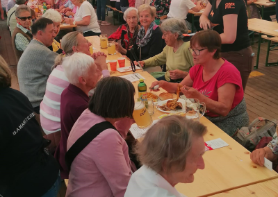 Innenansicht eines Festzelts mit langen Tischreihen, älteren Gästen beim Essen; Servierkräfte mit Schriftzug "OBER FIX"; Gläser mit Aufdruck "Zipfer"; Bühne mit Bergkulisse im Hintergrund.