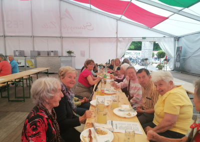 Mehrere ältere Personen sitzen an einer langen Holztischreihe in einem Festzelt und essen; Teller, Besteck und Gläser (auf einigen Gläsern die Aufschrift "Zipfer" sichtbar) auf dem Tisch; im Hintergrund Zelteingang und Fahrräder.