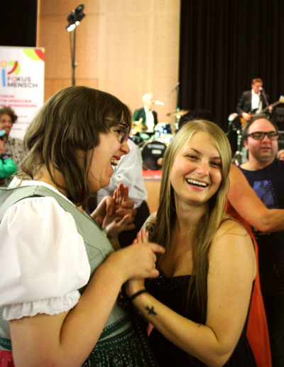 Zwei Frauen halten Händchen und lachen beim Tanzen vor einer Bühne; im Hintergrund Band und ein Banner mit 'FOKUS MENSCH'.