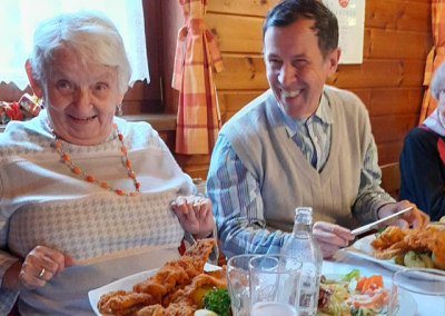 Zwei lächelnde ältere Personen sitzen an einem Tisch und essen gemeinsam; Teller mit Schnitzel, Beilagen und Getränken sichtbar.