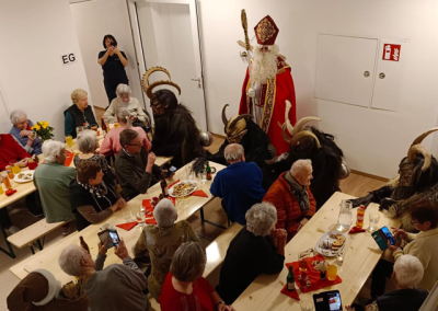 Ältere Teilnehmende an festlich gedeckten Tischen in einem Gemeinschaftsraum; eine als Nikolaus verkleidete Person steht neben mehreren in gehörnten, pelzigen Kostümen; Wandbeschilderung 'EG' sichtbar.