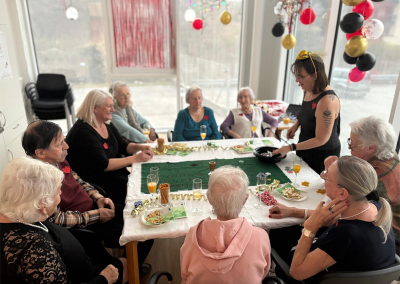 Innenraumaufnahme: mehrere ältere Personen sitzen an einem Tisch mit grünem Spieltuch, Spielmaterial, Getränken und Snacks; eine Frau steht und reicht Utensilien; hängende Dekorationen (Luftballons, Girlanden) sichtbar.