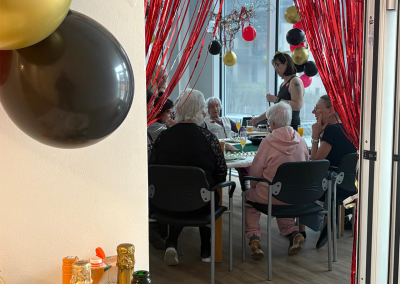 Eingang zu einem festlich geschmückten Raum mit handgemaltem Schild „Herzlich Willkommen im Casino“; im Inneren sitzen mehrere Gäste, im Vordergrund Champagnerflaschen.