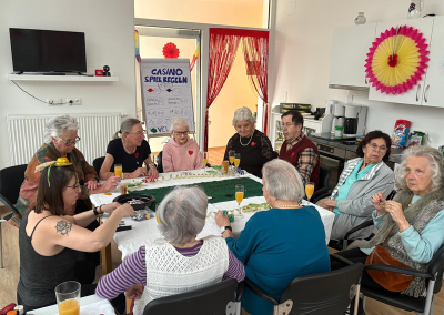Mehrere Personen sitzen an einem Tisch mit Spielutensilien, Getränken und Tischdekoration; im Hintergrund Flipchart mit der Überschrift 'CASINO SPIELREGELN'.