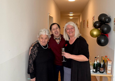 Drei Erwachsene stehen eng beieinander in einem Hausflur; rechts ein Tisch mit Sektflaschen und schwarzen/goldenen Luftballons.
