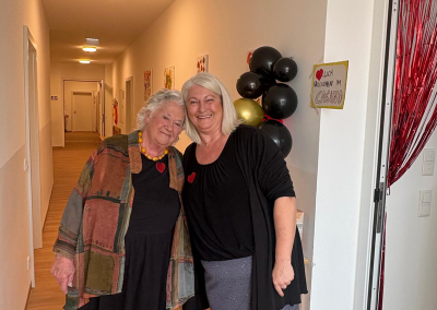 Zwei lächelnde Frauen in einem Flur vor schwarz‑goldenen Ballons und einem handgeschriebenen Schild mit dem Text "WILLKOMMEN IM CASINO".