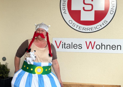 Person im Obelix-Kostüm mit roten Zöpfen und Plüschhund vor Wand mit Samariterbund-Logo und Schrift "Vitales Wohnen"