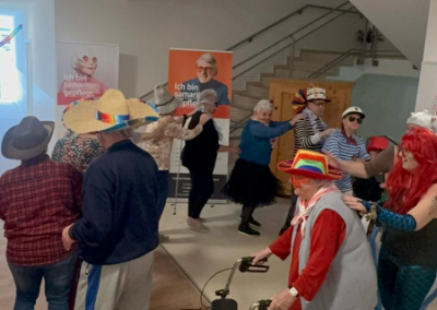 Gruppe von Menschen in bunten Kostümen und Hüten tanzt in einem Innenaufenthaltsbereich vor einer Treppe; ein Rollator im Vordergrund; im Hintergrund zwei Banner mit der Aufschrift 'Ich bin Samariter gepflegt'.