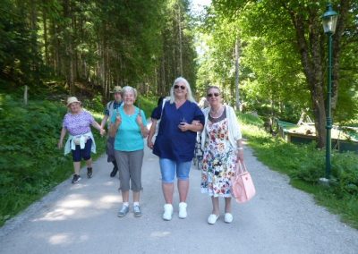 Drei Frauen Arm in Arm lächelnd auf einem breiten Waldweg, im Hintergrund weitere Personen; links und rechts hohe, grüne Bäume.
