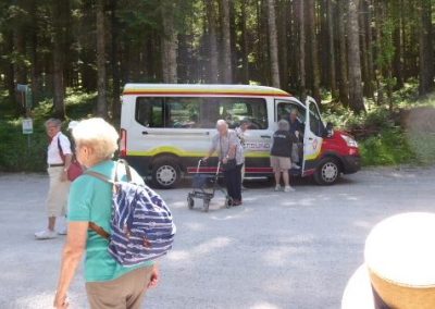 Ausflugsgruppe an einem Waldparkplatz, im Vordergrund Personen, im Hintergrund ein geparkter weißer Kleinbus mit farbigen Streifen; eine Person mit Rollator sichtbar