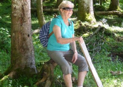 Frau mit Sonnenbrille sitzt auf einer herausragenden Baumwurzel im schattigen Nadelwald, trägt türkisfarbenes Oberteil, knielange Hosen, Rucksack und hält ein längliches Holzbrett.
