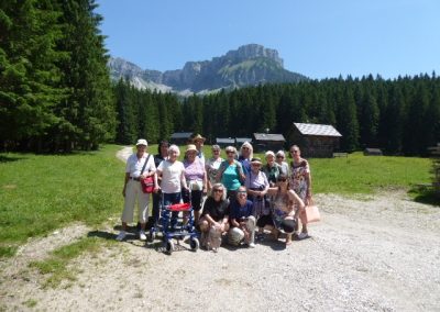 Gruppenfoto von etwa 15 Personen auf einer sonnigen Bergwiese vor Holzhütten, Nadelwald und markanter Felswand; vorn eine Gehhilfe sichtbar.