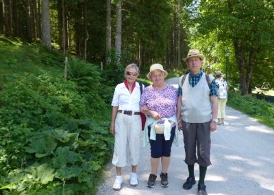 Drei Erwachsene in Sommerkleidung und Hüten stehen auf einem breiten, von Bäumen gesäumten Waldweg; eine weitere Person ist im Hintergrund sichtbar.