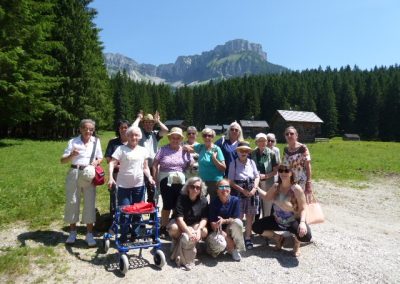 Gruppe von Personen posiert auf einer sonnigen Wiese vor Nadelwald und einer Felswand; Holzhütten im Hintergrund, ein Rollator vorn.