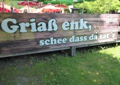 Großes Holzschild mit weißer Schrift: 'Griaß enk, schee dass da sat's'; davor Rasen, im Hintergrund Sonnenschirme und Sitzplätze.