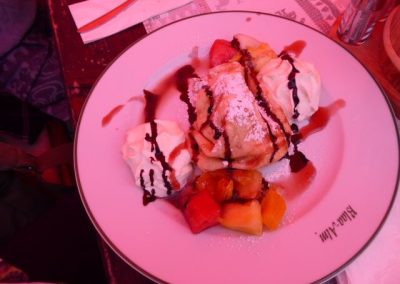 Nahaufnahme eines gefalteten Crêpe-ähnlichen Desserts auf weißem Teller mit Schlagsahne, geschnittenen Früchten, Schokoladensauce und Puderzucker; am Tellerrand ist teilweiser Text sichtbar.