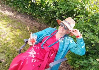 Frau mit Sonnenhut und Sonnenbrille sitzt im Garten vor einer grünen Hecke und hält ein Glas mit Getränk.