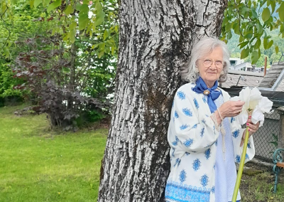 Ältere Frau lehnt an Baum und hält große weiße Blüte; Garten im Hintergrund.