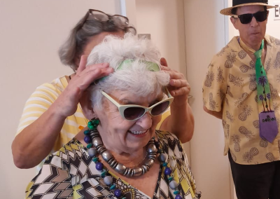 Ältere Frau mit Sonnenbrille und mehreren Halsketten, lächelnd, während eine Person ihr ein grünes Stirnband anpasst; im Hintergrund ein Mann mit Strohhut und dunkler Sonnenbrille.