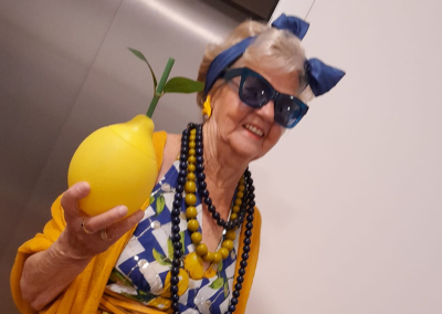Ältere Frau in blau-gelbem Kleid und gelbem Cardigan, mit dunkler Sonnenbrille und blauem Haarband, hält eine gelbe, zitrusförmige Deko-Zitrone.