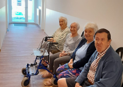 Vier ältere Personen sitzen in einer Reihe in einem hellen Innenflur; davor steht ein blauer Rollator.