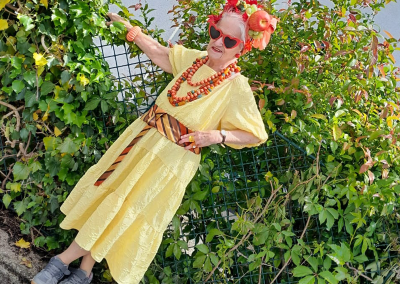 Ältere Frau im gelben Kleid mit roten herzförmigen Sonnenbrillen und orangenen Schmuck, lehnt an grünem Drahtzaun vor Efeu.