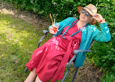 Frau in rotem Kleid und türkiser Jacke sitzt auf einem Gartenstuhl im Rasen, trägt Strohhut und Sonnenbrille und hält ein Getränk.