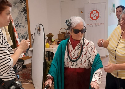 Ältere Frau mit Rollator und Sonnenbrille, von mehreren Personen begleitet; im Hintergrund Roll-up mit Schriftzug 'Samariterbund Bad Ischl'.
