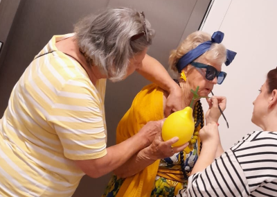 Ältere Frau in leuchtend gelbem Outfit und blauer Haarschleife mit dunkler Sonnenbrille, wird von zwei Helferinnen gestylt; eine hält ein großes gelbes Zitronen-Accessoire, eine trägt Lippenstift auf.