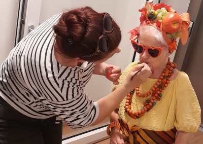 Ältere Frau im gelben Kleid mit künstlicher Obst- und Blumenkrone, roter Herz-Sonnenbrille und auffälligen Ketten, während ihr Lippenstift aufgetragen wird.