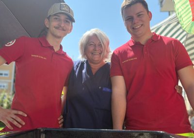 Drei Personen, zwei in roten Polohemden mit dem Schriftzug "SAMARITERBUND" und eine Frau in blauer Kleidung, lächeln in die Kamera hinter einem Grill mit Würsteln.