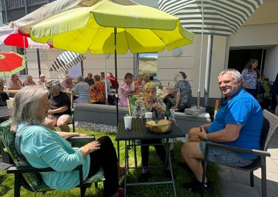 Drei Personen an einem Gartentisch unter Sonnenschirmen, im Hintergrund mehrere Gäste an langen Tischen; auf einem Schirm ist "Samariterbund" zu lesen.