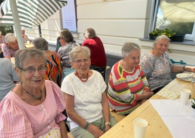 Vier ältere Frauen sitzen an einem gedeckten Holztisch im Freien; im Hintergrund weitere Teilnehmende, Sonnenschirme und ein Banner.